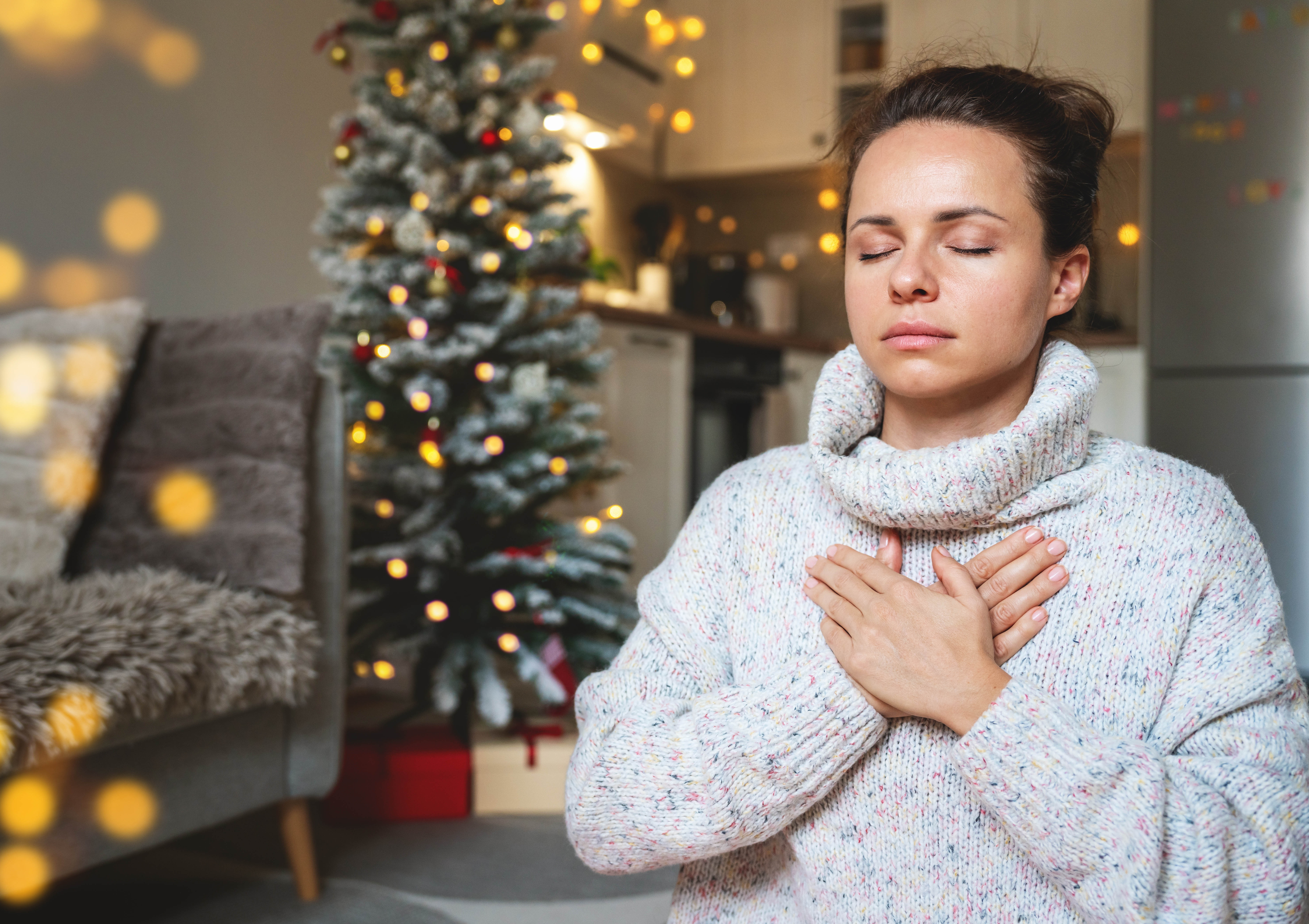 Woman with her eyes closed, hand on her chest, taking deep, relaxing breaths in front of the Christmas tree Woman with her eyes closed, hand on her chest, taking deep, relaxing breaths in front of the Christmas tree